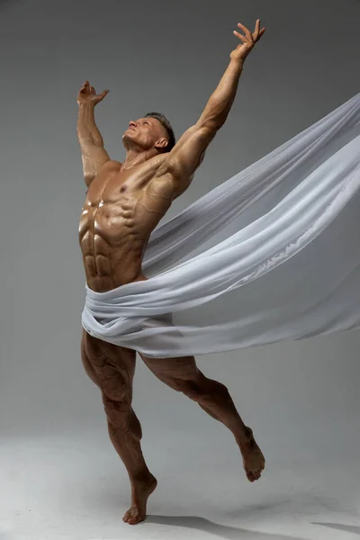 Aesthetics of the male athletic body. The male model pulls his hands up, standing on his toes. White fabric covers male muscular body