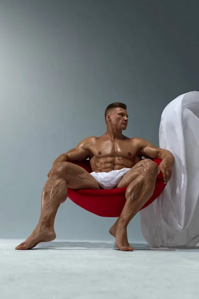 A muscular athlete with perfect abs poses while sitting in a red chair, covering himself with a white piece of fabric. The concept of 6 packs of perfect abs