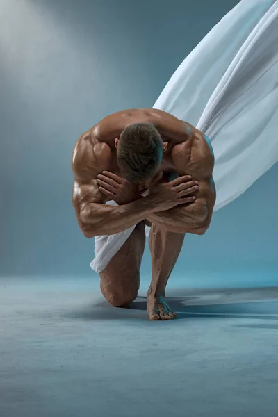 A strong, athletic man poses naked, covered with a white cloth, standing on one knee. Athletic muscular body with a low percentage of fat.
