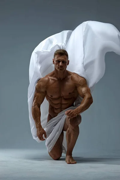 A strong, athletic man poses naked, covered with a white cloth, standing on one knee. Athletic muscular body with a low percentage of fat.