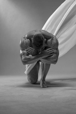 Güçlü, atletik bir adam çıplak poz veriyor, beyaz bir bez ile kaplı, tek dizinin üstünde duruyor. Düşük yağ yüzdesi olan atletik kaslı vücut. Siyah beyaz erkek fotoğrafı.