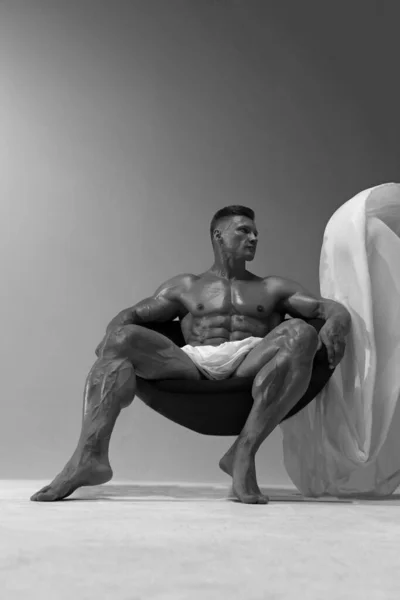 A muscular athlete with perfect abs poses while sitting in the chair, covering himself with a white piece of fabric. The concept of 6 packs of perfect abs. Black and white male photo