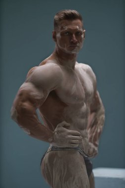 Mükemmel karın kasları olan güçlü kaslı bir adam stüdyoda boş bir arkaplanda poz veriyor. Atletik erkek vücudu antika bir heykel şeklinde beyaz kille kaplıdır.