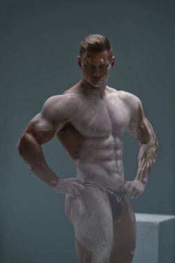 Mükemmel karın kasları olan güçlü kaslı bir adam stüdyoda boş bir arkaplanda poz veriyor. Atletik erkek vücudu antika bir heykel şeklinde beyaz kille kaplıdır.