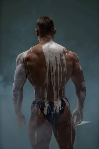 A strong man demonstrates a muscular relief back covered with white clay. Ideal body with a low percentage of fat as a result of a hard sports routine