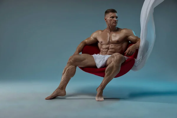 一位肌肉发达的运动员坐在红色的椅子上 用一块白色的面料盖住身体 完美地保持着腹肌的姿势 六包完美腹肌的概念 — 图库照片