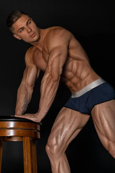 Bodybuilder man posing near chair at the studio in shorts, showing a perfect muscular body with a low percentage of fat. The concept of body aesthetics and a healthy lifestyle