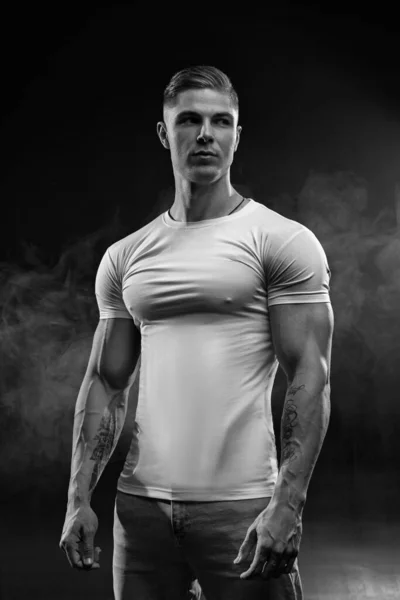 若いですセクシーな男性ボディビルダーの選手 スタジオの肖像画で黒ロフトで煙 男モデルで白Tシャツとジーンズで黒の背景黒と白の写真 — ストック写真