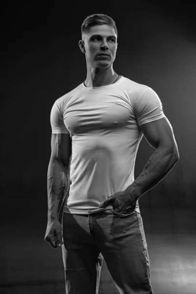 年轻性感的健美运动员 黑漆漆的工作室肖像画 身穿白色T恤的男模 手插牛仔裤口袋 背景黑白照片 — 图库照片