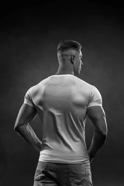 スタジオでポーズをとっている白いTシャツとジーンズを着た若い筋肉質の男 リアビュー 背景の黒と白の写真に空の黒のサイクロラマ — ストック写真