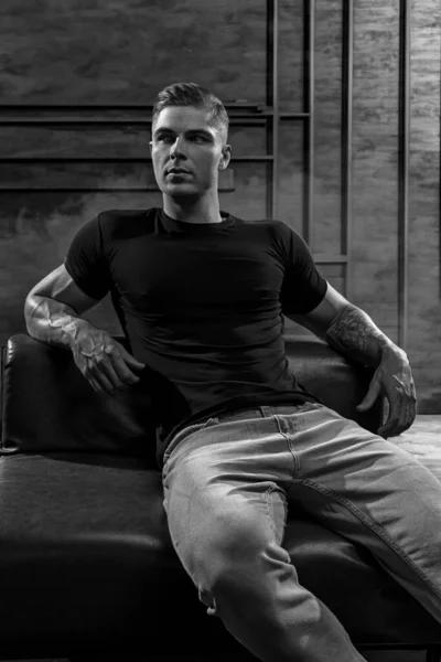 Genç seksi vücut geliştirme sporcusu deri koltukta otururken stilize edilmiş çatı katı tarzı bir duvarın arkasında poz veriyor. Siyah tişört ve kot pantolon içinde spor modeli, siyah ve beyaz fotoğraf