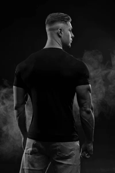 stock image Muscular model sports young man in jeans and black t-shirt on black background back view. Fashion portrait of brutal sporty healthy strong muscle guy black and white photo