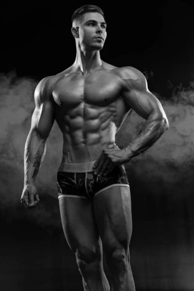 黒の背景にスタジオでショートパンツ姿の魅力的なセクシーボディビルダーは 筋肉の強い体を示しています ジムや身体の美学におけるハードワークの概念 — ストック写真
