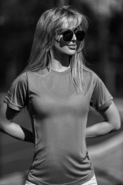 Sarışın atletik kız spor sahasında tişört, pantolon, beyaz spor ayakkabı ve siyah beyaz kulaklıkla duruyor.