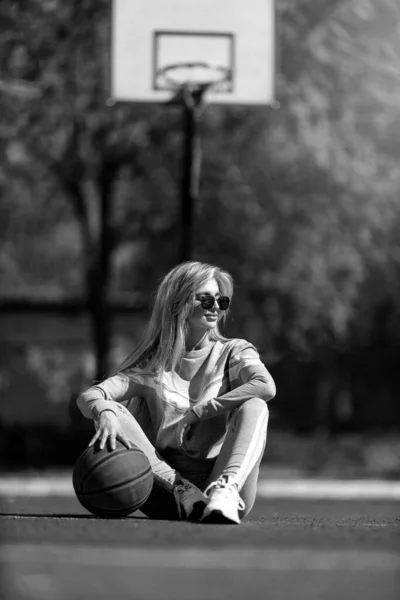 身穿灰色运动服 手里拿着一个黑白球的金发女运动员坐在篮球场上 — 图库照片