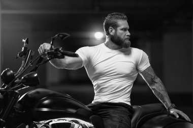 Seksi, yakışıklı, çekici, kaslı spor spor spor spor mankeni, beyaz dar tişörtlü motorcu, park yerinde, siyah beyaz bir fotoğrafta, vahşi, güzel bir motosikletin üzerinde oturuyor.