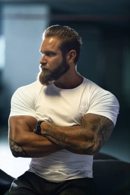 Seksi, yakışıklı, çekici, kaslı spor spor spor modeli, beyaz dar tişörtlü motorcu park yerinde vahşi, güzel bir motosikletin üzerinde oturuyor.