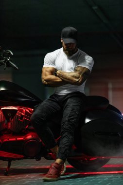 Seksi, yakışıklı, çekici, kaslı spor spor spor spor modeli, siyah şapkalı, beyaz dar tişörtlü motosiklet mankeni park yerinde vahşi, güzel bir motosikletin üzerinde oturuyor.