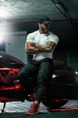 Seksi, yakışıklı, çekici, kaslı spor spor spor spor modeli, siyah şapkalı, beyaz dar tişörtlü motosiklet mankeni park yerinde vahşi, güzel bir motosikletin üzerinde oturuyor.