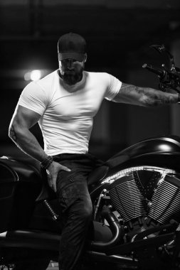 Seksi, yakışıklı, çekici, kaslı spor spor spor spor modeli siyah şapkalı, beyaz dar tişörtlü motosikletli siyah beyaz motosiklet park yerinde oturuyor.