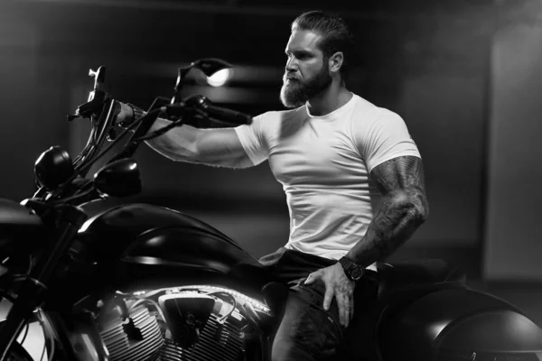 Seksi, yakışıklı, çekici, kaslı spor spor spor spor mankeni, beyaz dar tişörtlü motorcu, park yerinde, siyah beyaz bir fotoğrafta, vahşi, güzel bir motosikletin üzerinde oturuyor.