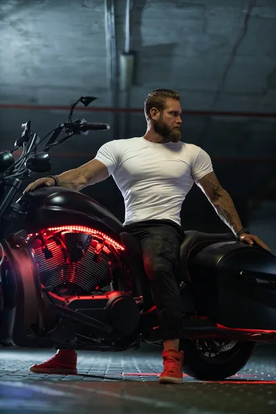 Seksi, yakışıklı, çekici, kaslı spor spor spor modeli, beyaz dar tişörtlü motorcu park yerinde vahşi, güzel bir motosikletin üzerinde oturuyor.