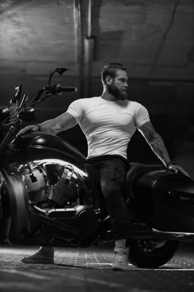 Seksi, yakışıklı, çekici, kaslı spor spor spor spor mankeni, beyaz dar tişörtlü motorcu, park yerinde, siyah beyaz bir fotoğrafta, vahşi, güzel bir motosikletin üzerinde oturuyor.