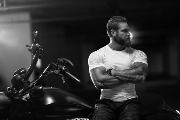 Seksi, yakışıklı, çekici, kaslı spor spor spor spor mankeni, beyaz dar tişörtlü motorcu, park yerinde, siyah beyaz bir fotoğrafta, vahşi, güzel bir motosikletin üzerinde oturuyor.