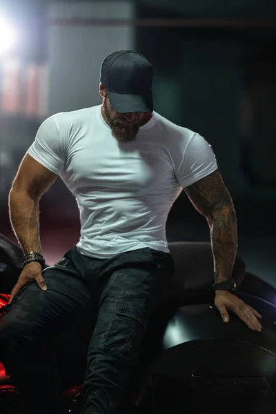 Seksi, yakışıklı, çekici, kaslı spor spor spor spor modeli, siyah şapkalı, beyaz dar tişörtlü motosiklet mankeni park yerinde vahşi, güzel bir motosikletin üzerinde oturuyor.