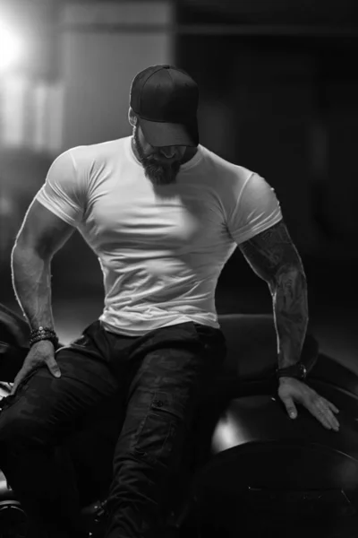 Seksi, yakışıklı, çekici, kaslı spor spor spor spor modeli siyah şapkalı, beyaz dar tişörtlü motosikletli siyah beyaz motosiklet park yerinde oturuyor.