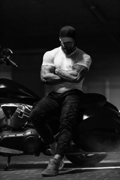 セクシーなハンサムな魅力的なスポーティな筋肉フィットネスモデル 黒い帽子と白のタイトなTシャツは駐車場 黒と白の写真で残酷な美しいオートバイに座っています — ストック写真