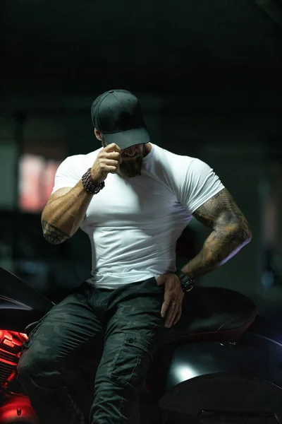 Seksi, yakışıklı, çekici, kaslı spor spor spor spor modeli, siyah şapkalı, beyaz dar tişörtlü motosiklet mankeni park yerinde vahşi, güzel bir motosikletin üzerinde oturuyor.