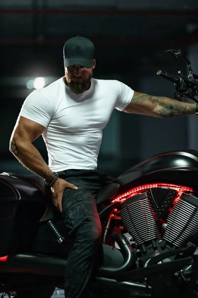 Seksi, yakışıklı, çekici, kaslı spor spor spor spor modeli, siyah şapkalı, beyaz dar tişörtlü motosiklet mankeni park yerinde vahşi, güzel bir motosikletin üzerinde oturuyor.