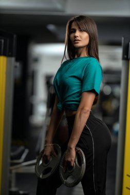 Yeşil tişörtlü, siyah eşofmanlı, elinde ağırlıklarla spor salonunda poz veren çekici esmer atletik kız. Sağlıklı yaşam tarzı, seksi kadın vücudu ve spor modası
