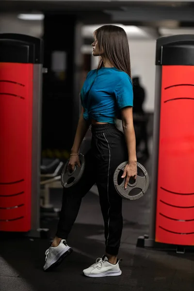 Attractive brunette athletic girl in blue T-shirt and black sweatpants posing with weights in her hands in the gym. Healthy lifestyle concept, sexy female body and sports fashion