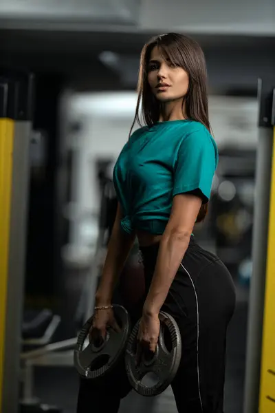 Attractive brunette athletic girl in green T-shirt and black sweatpants posing with weights in her hands in the gym. Healthy lifestyle concept, sexy female body and sports fashion