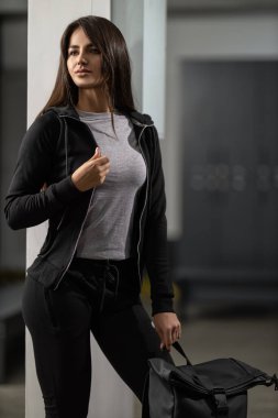 Güzel fitness modeli esmer, modern siyah eşofmanlı kadın spor salonunda siyah bir sırt çantasıyla poz veriyor.