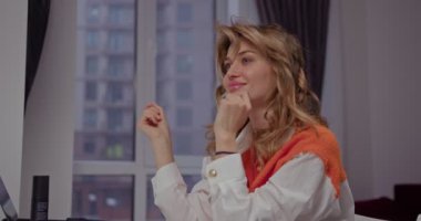 Beyaz gömlekli, parlak turuncu yelekli güzel bir kız şık kıvırcık saçlarına dokunuyor, neşeli saç stilini düzeltiyor.