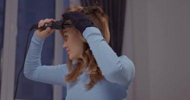 Güzel sarışın manken evde saç maşası yapıyor. Pencere kenarında saç maşası var.