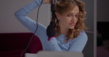 Sevimli sarışın kız evde aynanın önünde saç maşasıyla kıvırıyor..