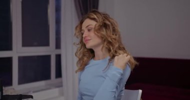 Genç ve güzel bir kız masada oturuyor ve aynanın önünde saçına dokunuyor.