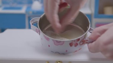Küçük bir çiçekli tencerenin içindeki malzemeleri ustalıkla karıştırıyor ve canlı renklerle dolu enfes bir minyatür mutfakta yemek pişirme sanatını sergiliyor..