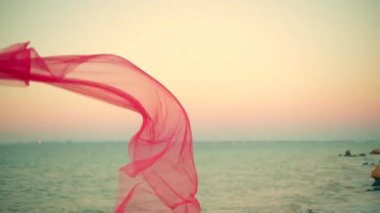 Uzun boylu kız gün doğumunda sahilde duruyor. Canlı kumaş parçasını tutarak, dingin ve büyüleyici atmosfer suyu yaratıyor.