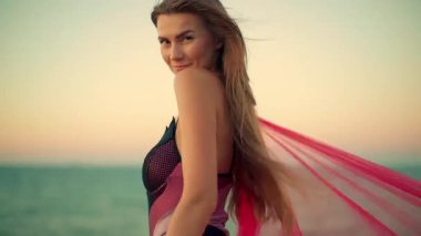 Gün doğumunda uzun boylu kız kumsalda duruyor yumuşak esinti uzun saçlarını ve renkli kumaşını dalgalandırıyor okyanusun ve gökyüzünün dingin zeminine neşe saçıyor
