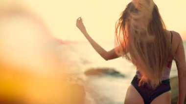 Uzun boylu kız kumlu kumsalda gün doğumunda uzun saçlarının ılık rüzgarda dalgalanmasına izin veriyor. Dalgaların ayaklarına hafifçe dokunması gibi sakin ve neşeli görünüyor.