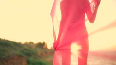 Güzel, uzun boylu bir kız kumlu sahilde gün doğumunda elinde süzülen kumaşlarla sabah rüzgarı altında dans ederek büyülü bir zemin oluşturur.