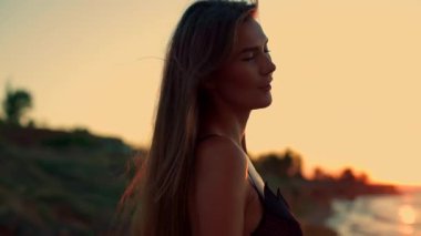 Uzun boylu kız plajda gün doğumunda zarif bir şekilde duruyor. Uzun saçları hafif esintide dalgalanan sakin dalgalar ve ılık altın ışık etrafında huzurlu bir atmosfer yaratıyor.