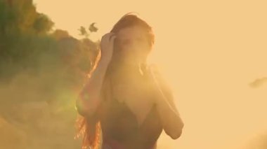 Uzun boylu bir kız kumsalda gün doğumunda saçlarının arasında parlıyor ve kumları aydınlatıyor. Huzurlu okyanus manzarasını ve taze sabah havasını seviyor.