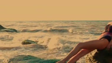 Uzun boylu kız, güneş yükselirken kayalık sahilde dinleniyor. Güneşin sıcacık parıltısı suya yansıyor. Huzurlu atmosfer yaratıyor.