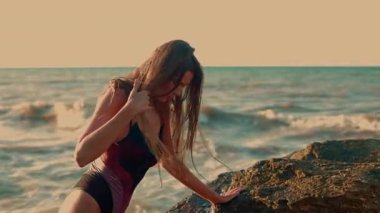 Uzun boylu kız sahilde güneşin doğuşunu seyrediyor yumuşak dalgalarla çevrili saçlarına dokunuyor ve zarif bir şekilde sabahın erken saatlerinde güneşlenirken dinlenme sahnesinin huzurunu yakalıyor.